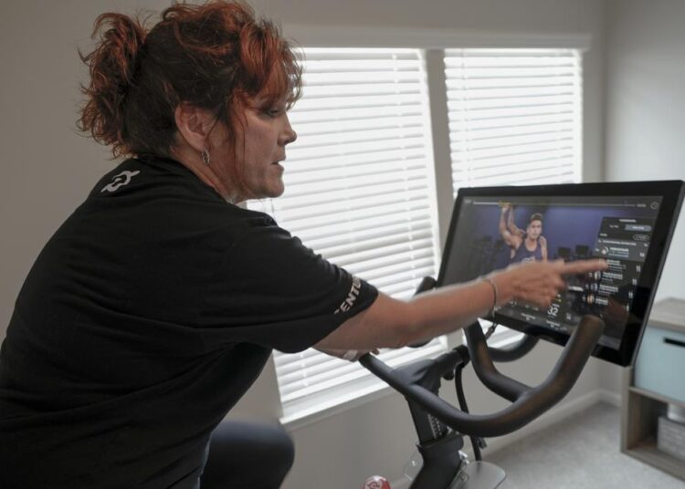 In this photo on Aug. 8, 2021, Cindy Cicchinelli, points out others on the screen listed to be cycling at the same time as she watches the online video while using her Peloton exercise machine in the workout room of her Pittsburgh townhouse.