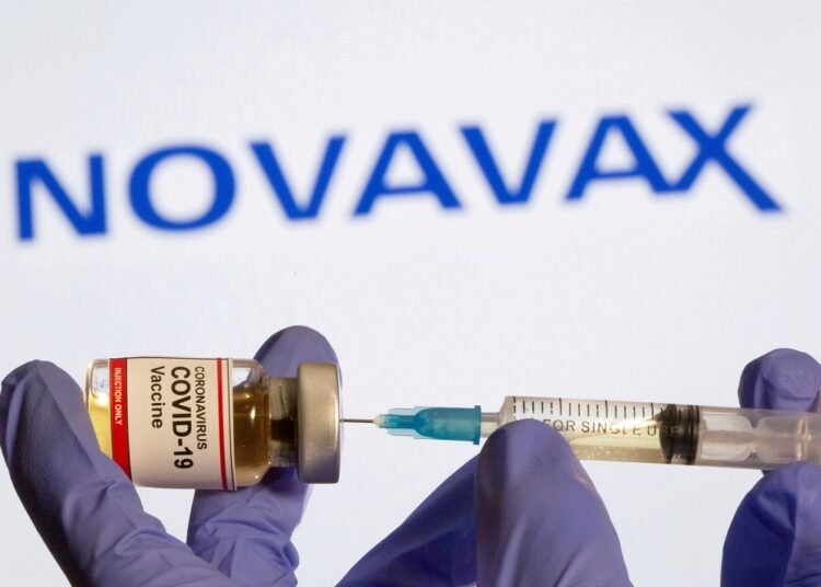 A woman holds a small bottle labeled with a "Coronavirus COVID-19 Vaccine" sticker and a medical syringe in front of displayed Novavax logo in this illustration taken, October 30, 2020.