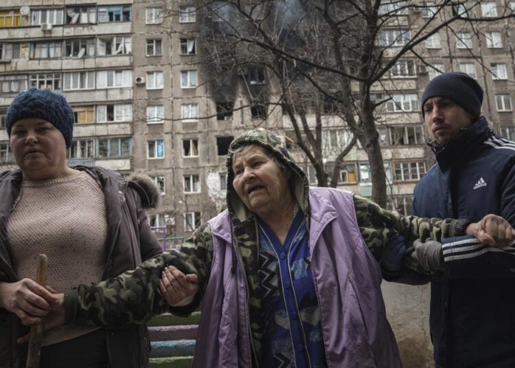 Sirens in Ukraine’s capital as civilians try to flee cities 1 - Egyptian Gazette People help an elderly woman to walk in a street with an apartment building hit by shelling in the background in Mariupol, Ukraine, Monday, March 7, 2022.
