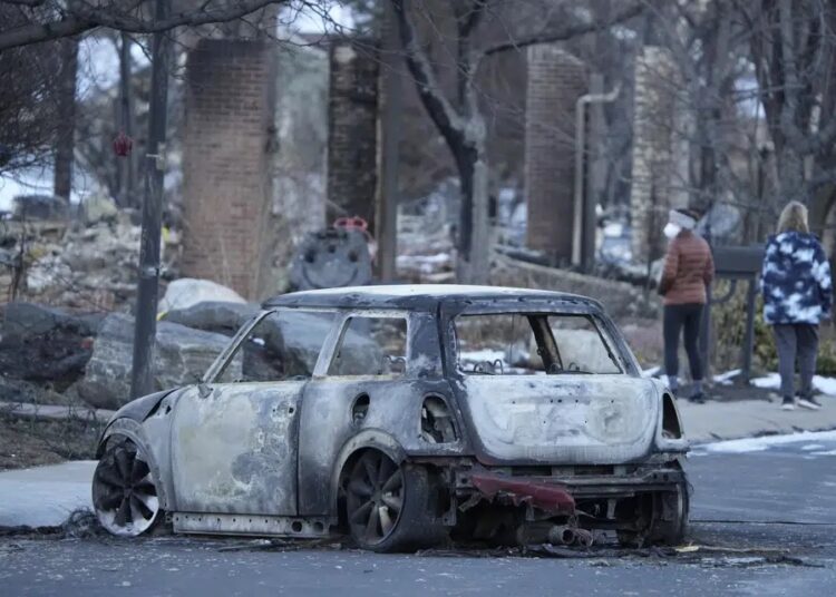 A charred Mini Cooper sits in Mulberry Street as pedestrians walk amid the rubble of homes destroyed by wildfires Tuesday, Jan. 4, 2022, in Louisville.
