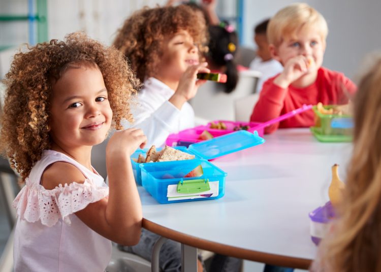 Here's How to Handle the End-of-Summer Scaries 1 - Egyptian Gazette Close up of smiling young children sitting at a table eating their packed lunches together at infant school, girl smiling to camera, selective focus